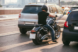Fototapeta Dmuchawce - rear view of a handsome biker dressed in a black T-shirt and black helmet jeans and sneakers. He is riding on cruiser motorcycle on background of 2 cars. Traffic stop