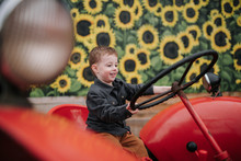 Cute Smiling Boy Holding Steering Wheel While Sitting On Red Tractor By Sunflower Farm