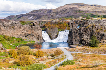 Hjalparfoss waterfall landscape of orange tree foliage in south Iceland in autumn fall season with water falling and highway road 32 and trail path hiking