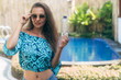 © romannoru - Portrait of brunette girl in sunglasses and blue top stands near pool and holds bottle of clean water in her hand