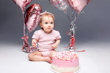 Little Girl's First Birthday In The Studio On A Gray Background Surrounded By Pink Balloons..Festive Portrait On The First Birthday Of A Little Girl In A Bodysuit With A Pink Cake And Balls.