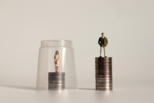 A Glass Ceiling Concept. A Miniature Man And A Miniature Woman Standing On A Pile Of Coins Of Different Heights.