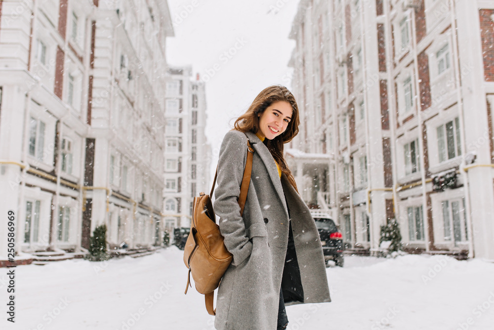 Stylish lady with brown backpack walking around city under snowfall. Outdoor photo of pretty girl with charming smile posing in gray coat on urban background in winter day. - obrazy, fototapety, plakaty 