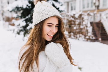 Wall Mural - Cozy winter portrait of fashionable joyful young woman with long brunette hair walking on street full with snow. Surprised true positive emotions, warm white woolen gloves, knitted hat