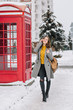 Full-length portrait of cheerful stylish lady talking on phone while walking around winter city. Outdoor photo of pretty caucasian female model with smartphone standing near red call-box.