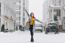 Full-length Portrait Of Inspired Female Model In Stylish Coat Posing With Pleasure In Winter City. Outdoor Photo Of Glad Blonde Woman Enjoying Snowfall During Walk Around Town.