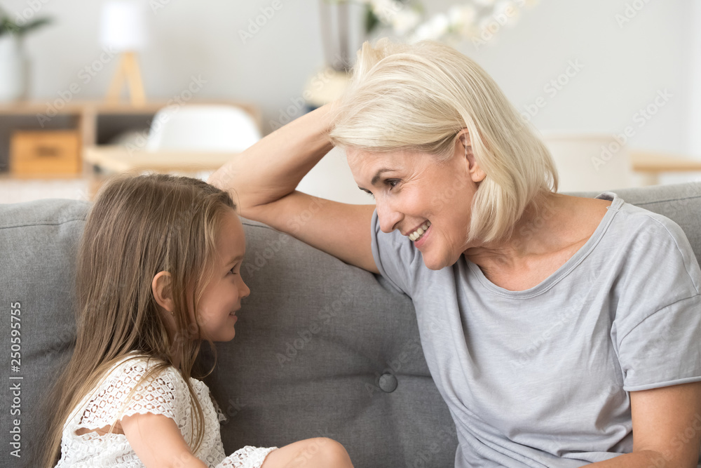 Little granddaughter talking with attentive grandmother, sitting on sofa - obrazy, fototapety, plakaty 