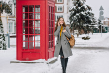 Amazing Young Woman In Gray Coat Talking On Phone On The Street With Green Spruce On Background. Outdoor Photo Of Glad Busy Girl With Brown Bag Walks Near Red Call-box.