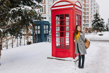 Full-length Portrait Of Lovely European Lady With Leather Bag Standing Near Phone Booth And Looking Away. Outdoor Photo Of Stunning White Woman In Gray Coat Posing Next To Call-box In Winter Day.