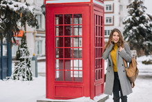 Stunning Brunette Woman In Yellow Cardigan Standing Near British Call-box In Winter Day. Outdoor Photo Of Adorable Girl In Trendy Coat Posing Beside Phone Booth With Spruce On Background..