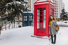 Outdoor Portrait Of Fashionable Brunette Girl With Backpack Posing Beside Red Call-box During Weekend In England. Full-length Photo Of Refined Woman In Gray Coat Smiling Near Phone Booth In Winter..