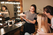 © estradaanton - Young professional make up artists with client in room. Woman on left hold eyeshadows palette and brush. Blonde and bruette women look in mirror together.