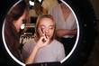 © estradaanton - Look in mirror in beauty room. Make up artist put mascara on eyelashes. Young blonde woman sit on chair with closed eyes. Hardresser work behind client.