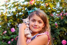 Portrait Of Pretty Young Girl Playing With Cute Little Kitten In Garden With Pink Blossom Roses In Sunny Spring Day