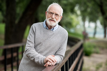 selective focus of senior man in grey pullover and glasses standing by wooden bridge railing and looking at camera