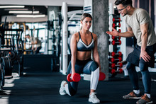 Young Muscular Woman Doing Weighted Lunge With Dumbbells, With Personal Trainer Motivating Her.