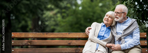 Fotomural smiling senior couple embracing while sitting on wooden bench in park
