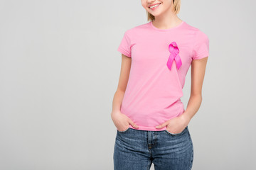 cropped view of smiling young woman in pink t-shirt with breast cancer awareness ribbon, isolated on grey