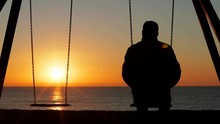 Back View Silhouette Of A Man Alone Contemplating Sunset Sitting On A Swing On The Beach