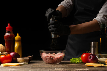 The chef peppers the fresh ground beef for the patties in the burger. On the background are the ingredients for the burger. Horizontal photo. Cooking delicious food