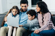 © LIGHTFIELD STUDIOS - smiling hispanic family sitting on sofa and looking at laptop at home