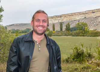 Wall Mural - Handsome young blond man is smiling against of white cliffs of Mount Ak-Kaya. Tourist wearing black leather jacket, in left ear hands fries. Theme for traveling to sights of Crimea.