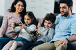 © LIGHTFIELD STUDIOS - cheerful parents sitting on sofa near kids playing video game at home