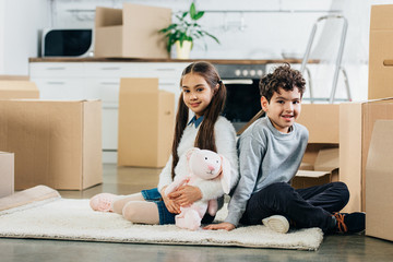 happy kids sitting on carpet with soft toy near boxes while moving in new home