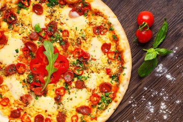 Close up Italian pizza with melted cheese, sausage, snack sticks, pepper and fresh green oregano leaves on a brown table decorated by mushrooms, red sweet pepper and cherry tomatoes