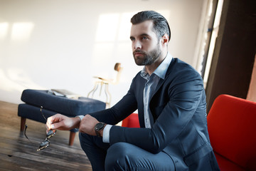 Wall Mural - Feeling confident. Attractive and stylish businessman in a fashion suit is looking away while sitting on office sofa