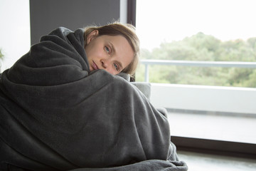 Sick woman sitting on sofa, embracing knees, looking at camera