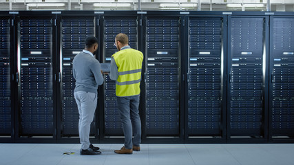 Poster - Bearded IT Specialist in Glasses and High Visibility Vest with a Laptop Computer and Black Technician Colleague Talking in Data Center while Standing Next to Server Racks. Running Maintenance. 