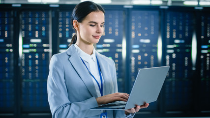 female it specialist is working on laptop in data center next to server racks.