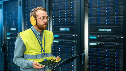 Aufkleber - Bearded IT Specialist in Glasses and Headphones, wearing High Visibility Vest is Working on Laptop in Data Center Next to Server Racks. Running Diagnostics or Doing Maintenance Work.