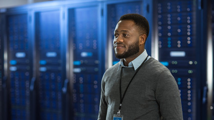 Aufkleber - Black Data Center IT Technician Standing in the Middle of a Server Rack Corridor. He is Smiling.