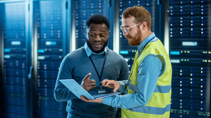 Poster - Bearded IT Specialist in Glasses and High Visibility Vest with a Laptop Computer and Black Technician Colleague Talking in Data Center while Standing Next to Server Racks. Running Diagnostics. 