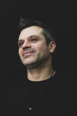 Wall Mural - A gay man in black t shirt on a black background - wearing pride necklace, with salt and pepper hair and stubble. 