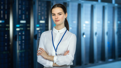 Poster - Female IT Specialist is standing at the Camera in Data Center Next to Server Racks and Looking at the Camera. Her Arms Crossed and She Smiles to the Camera.