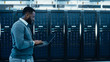 © Gorodenkoff - Black Data Center IT Technician Walking Through Server Rack Corridor with a Laptop Computer. He is Visually Inspecting Working Server Cabinets.