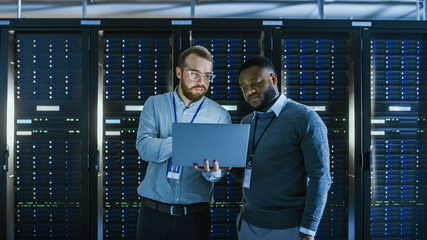 Bearded IT Technician in Glasses with a Laptop Computer and Black Male Engineer Colleague are Talking in Data Center while Working Next to Server Racks. Running Diagnostics or Doing Maintenance Work. 