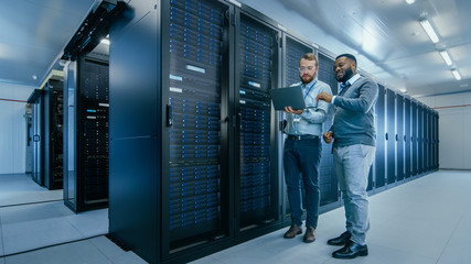 Bearded IT Technician in Glasses with a Laptop Computer and Black Male Engineer Colleague are Talking in Data Center while Working Next to Server Racks. Running Diagnostics or Doing Maintenance Work. 