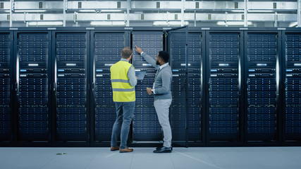 Aufkleber - Bearded IT Engineer in Glasses and High Visibility Vest with a Laptop Computer and Black Technician Colleague Talking in Data Center while Standing Next to Server Racks. Running Maintenance. 