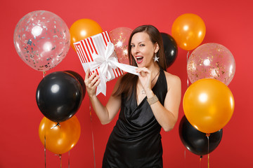 Cunning young girl in black dress celebrating opening red box with gift present on bright red background air balloons. International Women's Day, Happy New Year, birthday mockup holiday party concept.