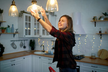 Young Happy Woman Twists A Light Bulb In The Kitchen Lamp