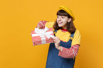 Pretty girl teenager in french beret, denim sundress looking aside, holding red striped present box with gift ribbon isolated on yellow background. People emotions, lifestyle birthday holiday concept.