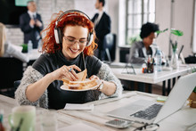 Happy redhead businesswoman eating a sandwich at her office desk.