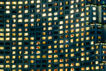 Frontal view of the night facade of building with a lot of windows and Lamp in many room within hotel.