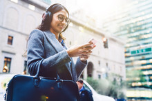 Smiling Indian Female Listening To Music In Headphones While Standing On City Street And Relaxing After Work. Young Woman Enjoying Playlist In Earphones. Watching Content In Social Network, Millennial