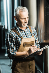 professional male brewer in working overalls writing in notepad in brewery