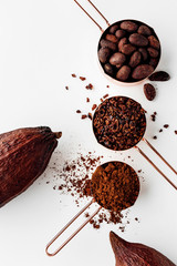 Rose gold measuring cups of cocoa beans, cacao nips, cocoa powder and cocoa pods on a white background, healthy food concept